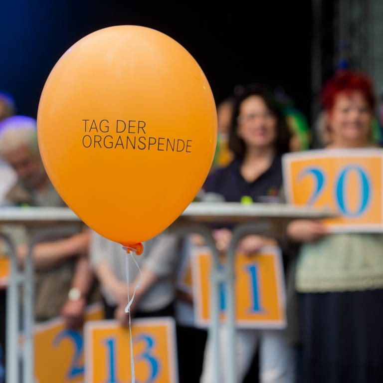 Ein Luftballon mit der Aufschrift "Tag der Organspende". Im Hintergrund Menschen, die Schilde hochalten, auf denen die Anzahl der Jahre steht, die sie nun mit Spenderorgan leben.