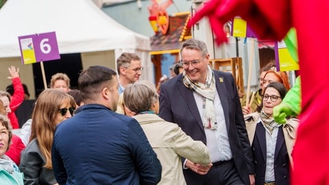 RLP-Tag: Ministerpräsident Schweitzer und seine Ehefrau begrüßen als Teil einer bunten Parade aus Fuss- und Musikgruppen die Zuschauer. (Foto: picture-alliance / Reportdienste, picture alliance/dpa | Andreas Arnold) RLP-Tag: Ministerpräsident Schweitzer und seine Ehefrau begrüßen als Teil einer bunten Parade aus Fuss- und Musikgruppen die Zuschauer.