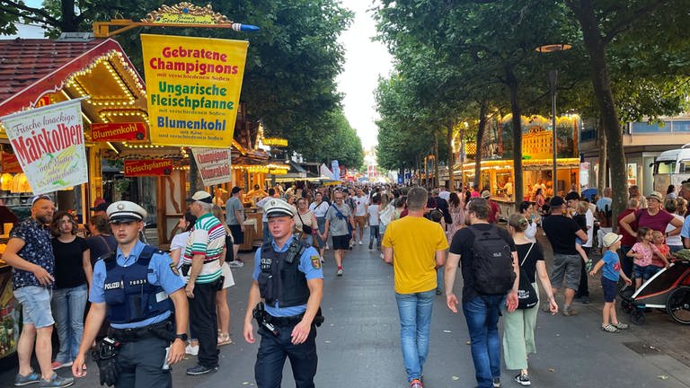 Zwei Streifenpolizisten laufen beim Johannisfest in Mainz am Rhein entlang. Dort stehen viele Buden, an denen Essen und Getränke gekauft werden können. 
