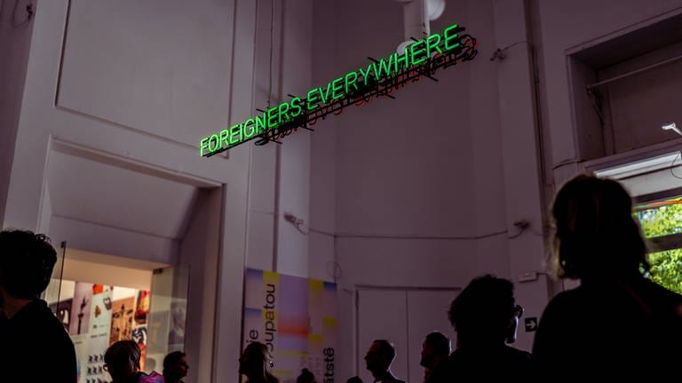 Besucher in einem dunklen Raum mit der Neon-Schrift: "Foreigners Everywhere"