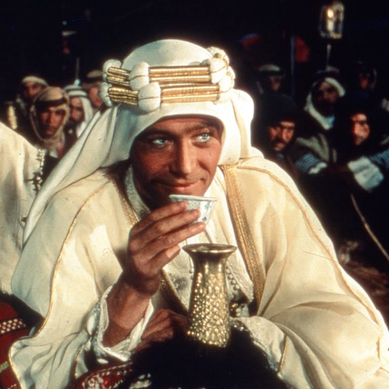 Moviestill von Lawrence von Arabien. Lawrence (Peter O'Toole) sitzt im arabischen Outfit vorne im Bild und trinkt Tee.