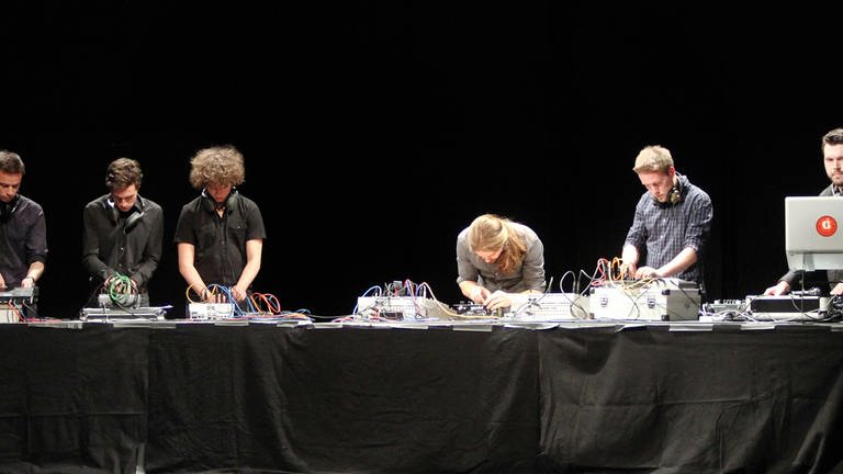 sechs junge Männer stehen an einem langen Tisch und beugen sich über elektronische Geräte