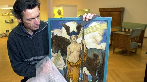 Mann packt eine Keramiktafel im Jugendstil aus, auf der eine „Minerva" abgebildet ist. (Foto: picture-alliance / Reportdienste, Rolf Haid) Mann packt eine Keramiktafel im Jugendstil aus, auf der eine „Minerva" abgebildet ist.