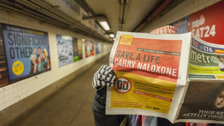 Ein Pendler liest 2017 eine Zeitung mit einer Anzeige des New Yorker Gesundheitsministeriums: Menschen werden aufgefordert, Naloxon bei sich zu haben – ein Notfallmedikament für eine Opioid-Überdosis. Während sich eine Opioid-Epidemie im ganzen Land ausbreitet, fordern Gesundheitsbehörden das Mitführen von Naloxon für den Fall, dass man auf einen überdosierten Süchtigen trifft.
