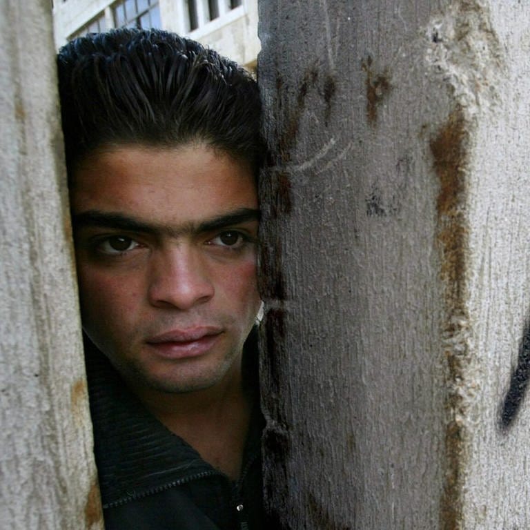 Ein Palästinenser, der auf der Westjordanlandseite des „Trennzauns“ in Abu Dis steht, schaut im November 2003 durch eine Lücke, um zu sehen, ob israelische Polizisten in der Nähe auf der Ost-Jerusalem-Seite der Mauer sind. Auf der Mauer steht "Apartheid". (Foto: dpa Bildfunk, picture-alliance / dpa/dpaweb | Pavel_Wolberg)