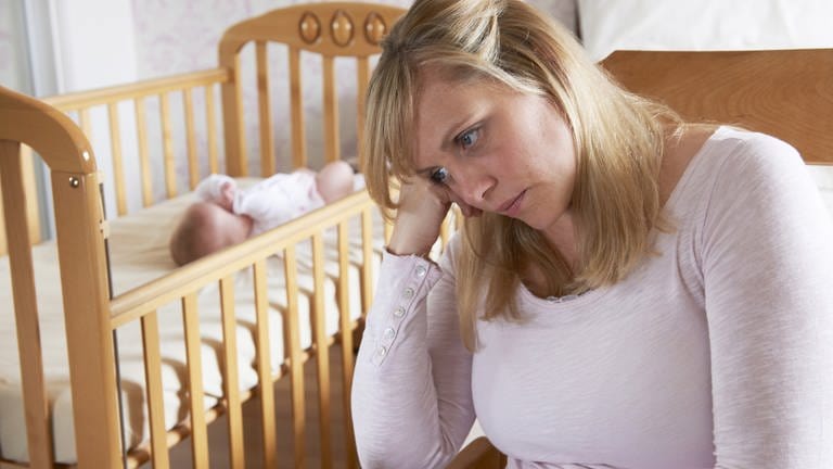 Mutter sitzt traurig vor einem Bettchen, in dem das Baby schläft: Nach der Geburt erleben viele Mütter einen Babyblues. Bleibt dieses Gefühl, haben sie eine Wochenbettdepression. Die Erkrankung kann jede Mutter treffen, bleibt aber oft unerkannt.