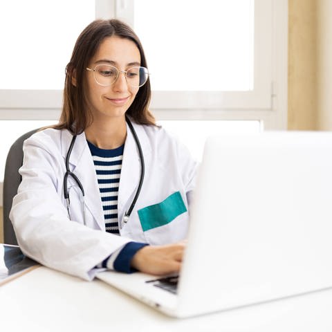 Junge Ärztin in Arztuniform und Brille sitzt mit Laptop am Tisch