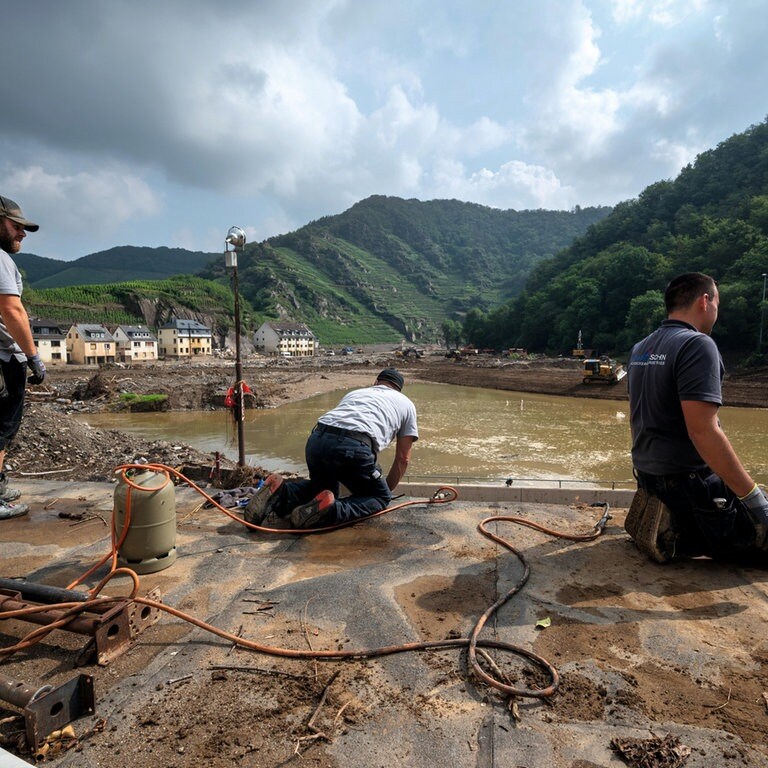 Nach der Flutkatastrophe in RLP: drei Handwerker bei Aufräumarbeiten an der Ahr. Im Hintergrund fließt der Fluss und sind Weinberge zu sehen. (Foto: SWR, Harald Tittel, picture alliance/dpa)