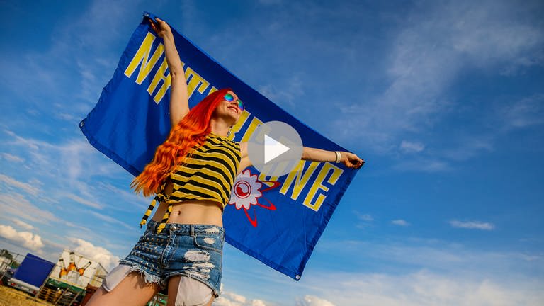 Eine Besucherin mit langen roten Haaren und kurzem gelb-schwaz geringelten Top und Shorts auf dem Festivalgelände mit sommerlichem blauen Himmel. Sie hält hinter sich eine blaue Fahne mit gelbem Aufdruck "Nature One" (Foto: SWR, Thomas Niedermueller)