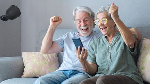 Zwei Senioren jubeln beim Blick auf ein Handy: Die vorherrschende Erklärung besagt, dass das Glück im Alter zunimmt, doch Studien zeigen einen gravierenden Fehler auf