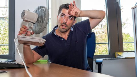 Arbeiter in Büro kämpft mit Ventilator gegen sommerliche Temperaturen Hitze im Büro
