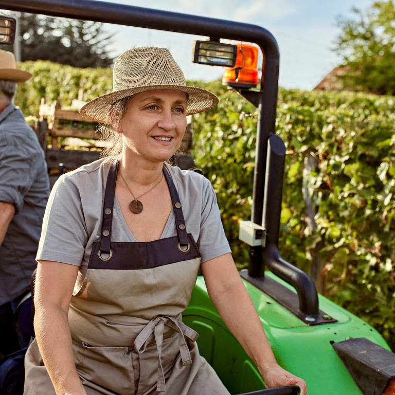 Eine lächelnde Frau fährt einen Traktor durch auf einem Acker, hinter ihr sitzt ein Kollege